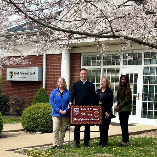 Four people stand holding a "City of Murray Beautification Award" sign in front of First Financial Bank, with blooming cherry blossom trees framing the scene.