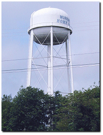 A ground-level picture looking up at the white-painted water tower for the city of Murray. The water tower is drastically taller than the trees below.