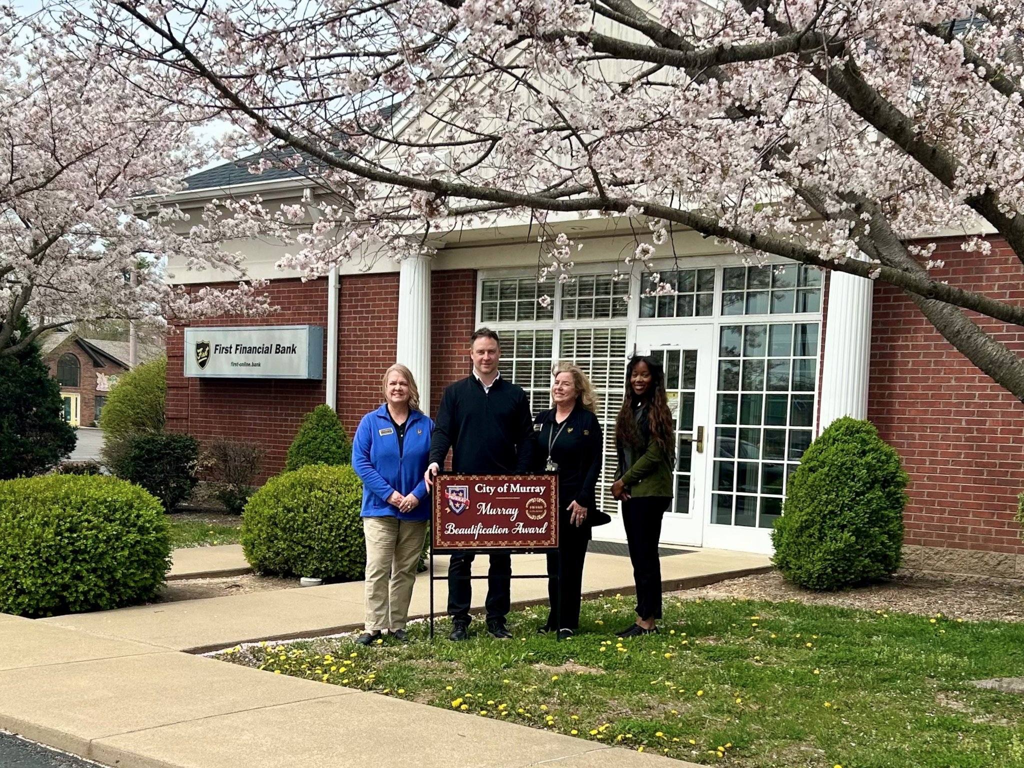 Four people stand holding a 'City of Murray Beautification Award' sign in front of First Financial Bank, with blooming cherry blossom trees framing the scene.