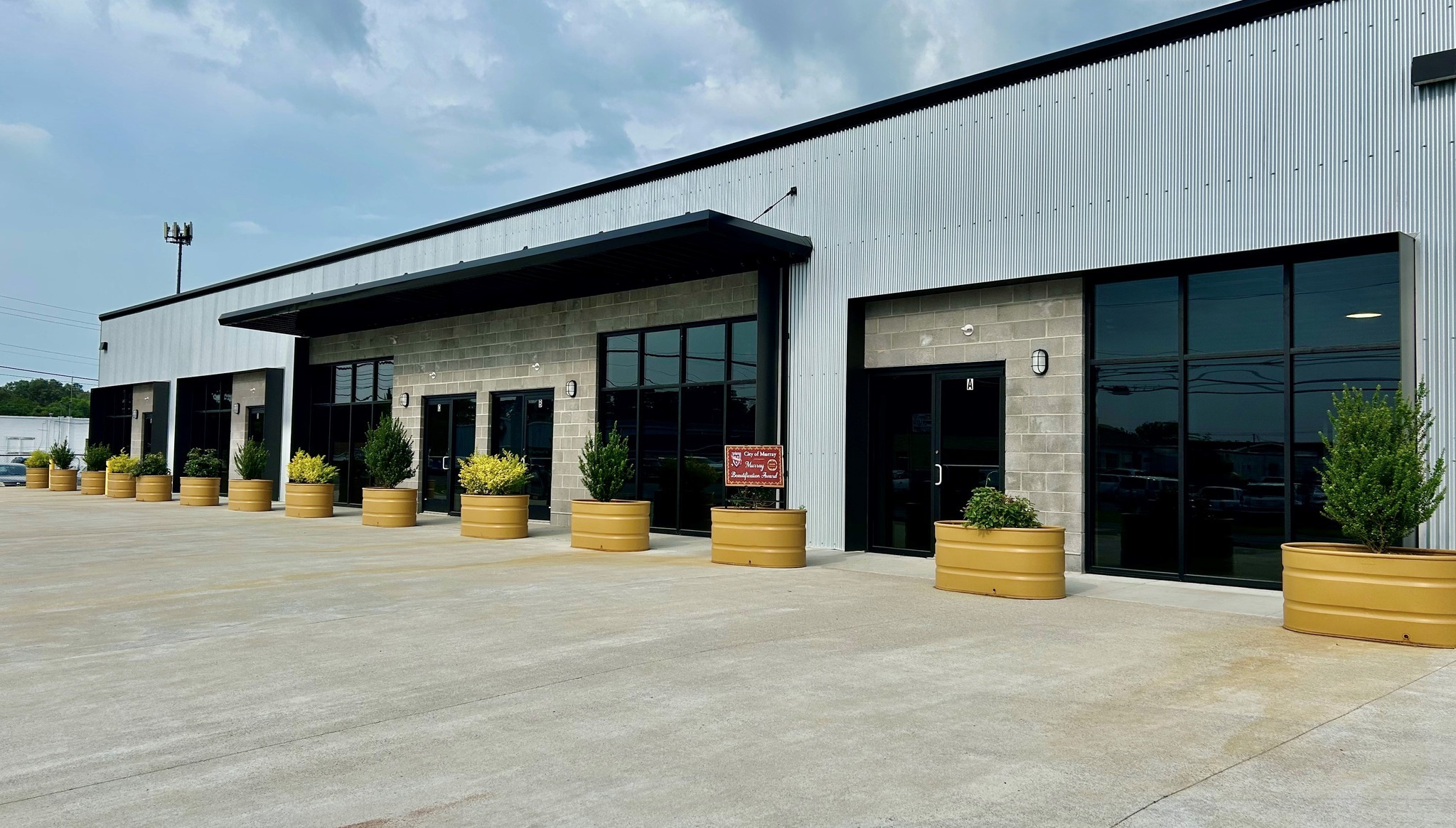 Modern commercial building with large windows and a row of yellow planters, displaying a 'City of Murray Beautification Award' sign.