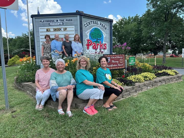 A group of nine people pose in front of a flower bed and sign for Central Park and Playhouse in the Park in Murray, Kentucky, with a 'City of Murray Beautification Award' sign also visible.