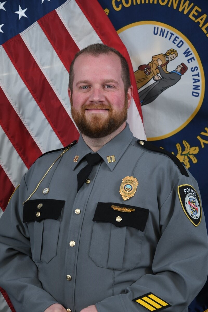Image of police officer in gray uniform in front of the United States and State of Kentucky flags.