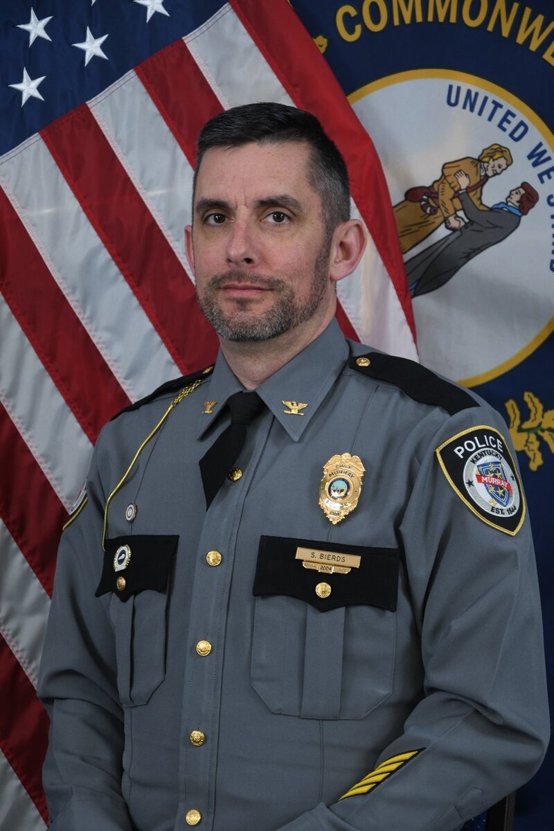 Police officer in gray uniform with badge and rank insignia standing before U.S. and Kentucky flags.