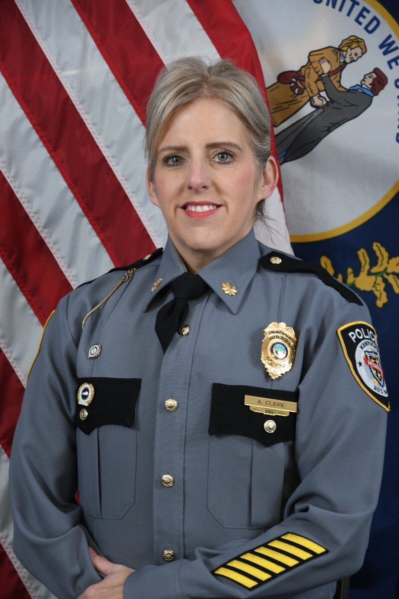 Police officer in gray uniform with badge and insignia standing before U.S. and Kentucky flags.