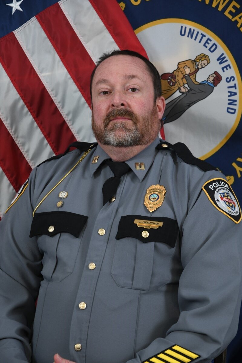 Police officer in gray uniform with badge and rank insignia standing before U.S. and Kentucky flags.