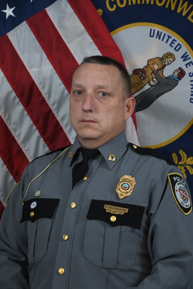 Police officer in gray uniform with badge and rank insignia standing before U.S. and Kentucky flags.