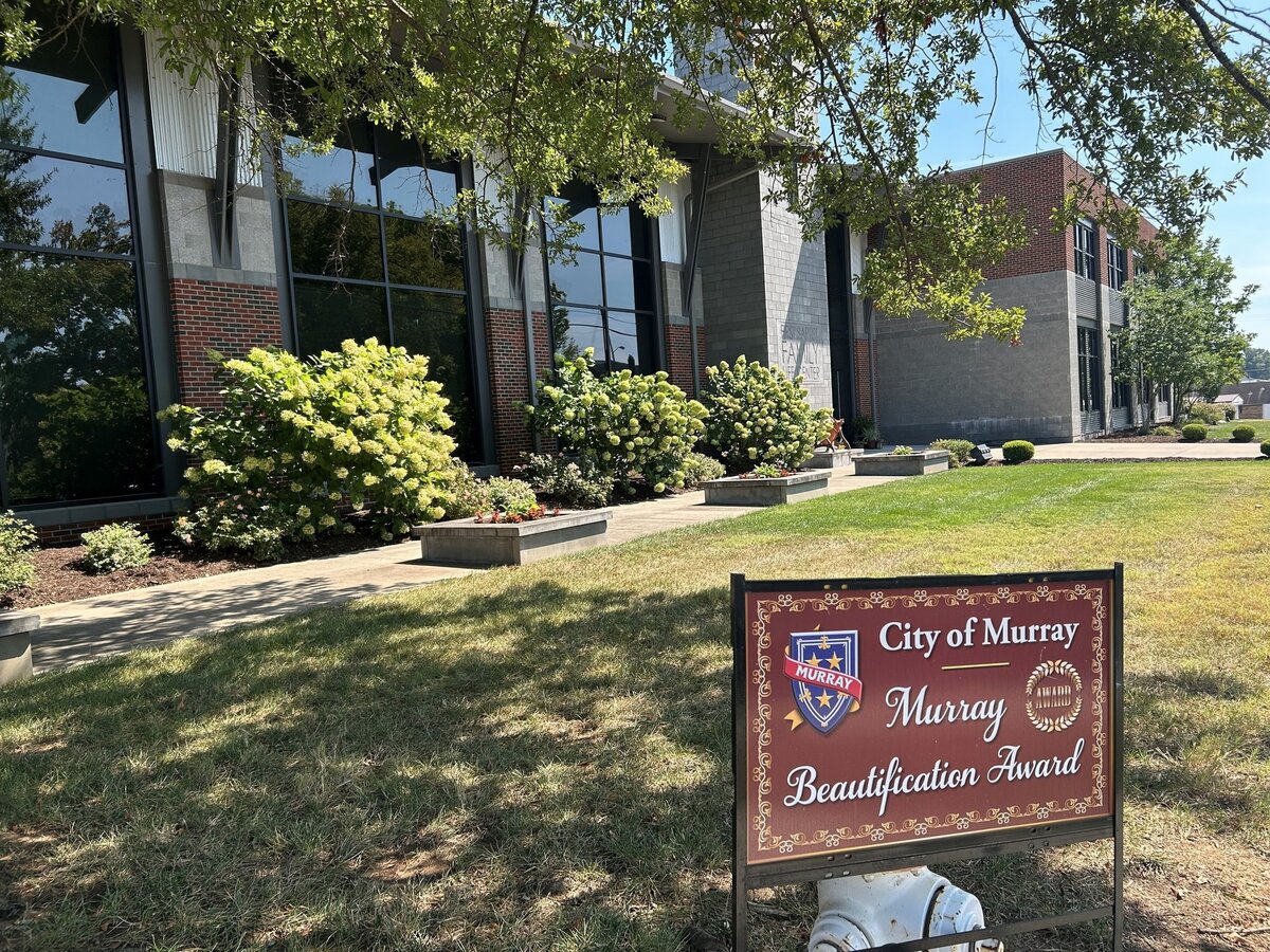 A modern brick and glass building is bordered by green shrubs and flowering plants. In the foreground, a brown sign reads ‘City of Murray, Murray Beautification Award’ with the city crest. Trees cast shade over the grassy area on a sunny day.