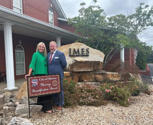 A man and woman stand smiling beside a brown ‘City of Murray, Murray Beautification Award’ sign in front of a red-brick building with white columns. Behind them is a large engraved stone reading ‘IMES Funeral Home & Crematory,’ surrounded by decorative rocks and landscaped plants.